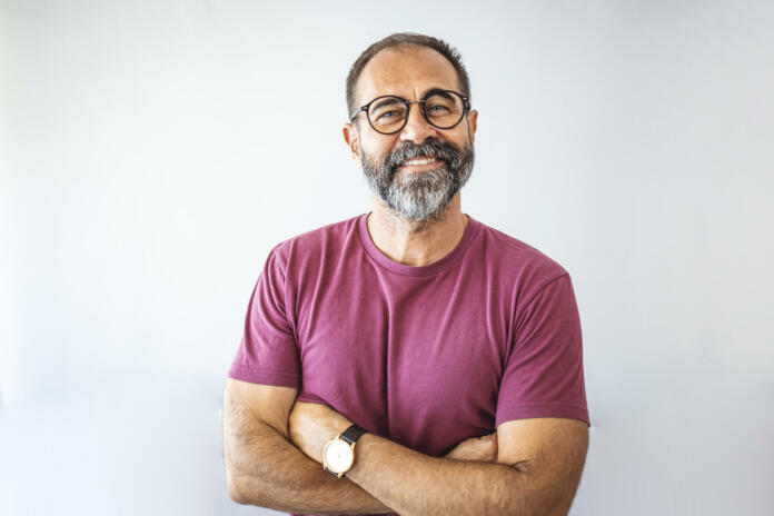 Portrait of mature businessman. Male executive is wearing T-shirt. Professional is smiling against gray background. Confident businessman with arms crossed. Mature mixed race man smiling