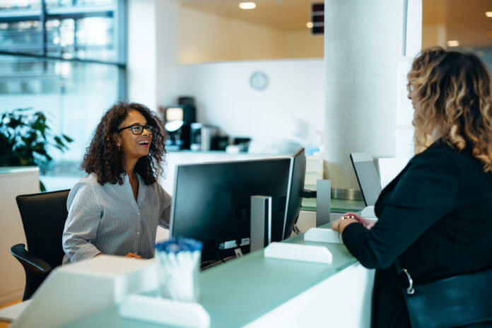 Receptionist assisting a woman standing at front desk. Woman standing at the reception talking with a friendly receptionist behind the desk of municipality office.