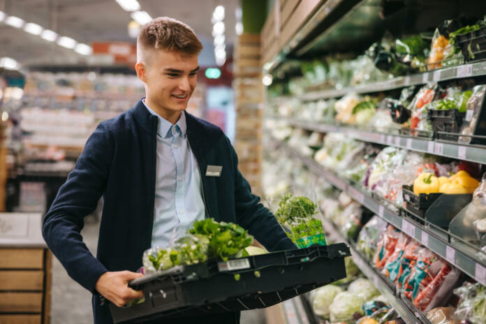 Shop assistant in supermarket re-stocking fresh vegetables in shelves of produce section.