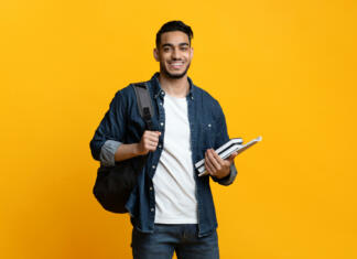 Študent Tomi Smart arab guy student with backpack and bunch of books smiling at camera, copy space for advertisement over yellow studio background. Education, university, college, studying, course concept