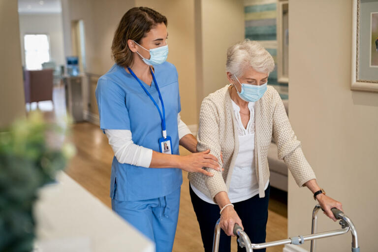 Smiling nurse with face mask helping senior woman to walk around the nursing home with walker. Young lovely nurse helping old woman with surgical mask for safety against covid-19 using a walking frame in hospital corridor. Friendly caregiver and elderly patient walking in hallway wearing protective face mask during covid pandemic.