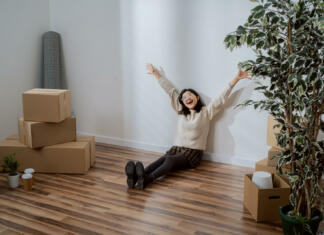 Kako do postelje v študentu? Smiling young woman of Asian Japanese beauty sits against wall in new apartment dressed in sweater and skirt, she is happy about move raises hands up in happiness next to unpacked boxes of stuff