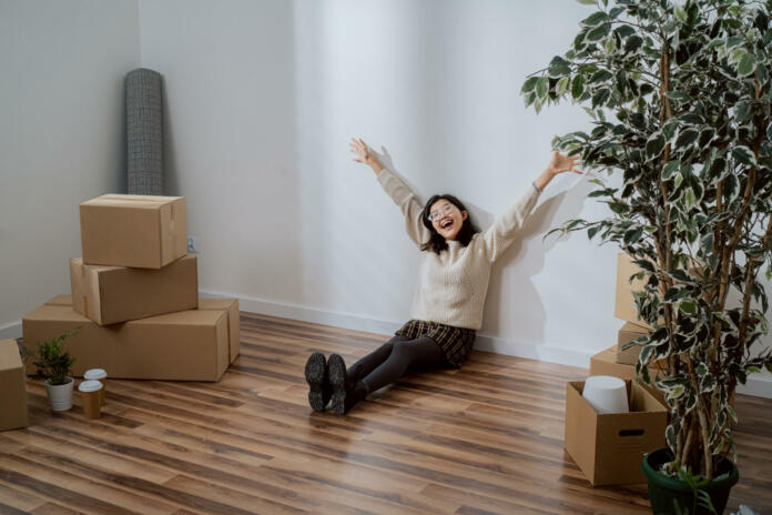 Smiling young woman of Asian Japanese beauty sits against wall in new apartment dressed in sweater and skirt, she is happy about move raises hands up in happiness next to unpacked boxes of stuff