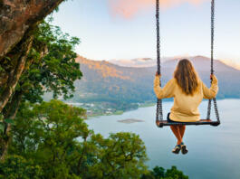 Summer vacation. Young woman sit on tree rope swing on high cliff above tropical lake. Happy girl looking at amazing jungle view. Buyan lake is popular travel destinations in Bali island, Indonesia