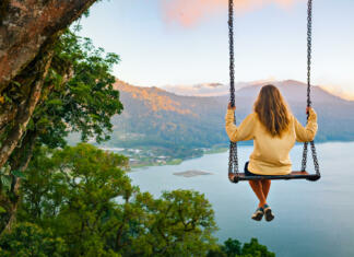 Izlet, ki je mnogo več kot to Summer vacation. Young woman sit on tree rope swing on high cliff above tropical lake. Happy girl looking at amazing jungle view. Buyan lake is popular travel destinations in Bali island, Indonesia