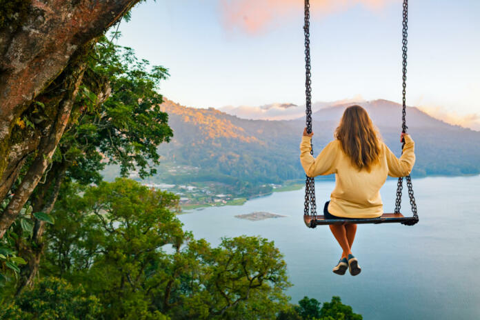 Summer vacation. Young woman sit on tree rope swing on high cliff above tropical lake. Happy girl looking at amazing jungle view. Buyan lake is popular travel destinations in Bali island, Indonesia