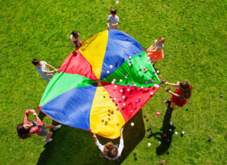 Podarjamo 10 Drinkopoly-ev Top view picture of kids standing in a circle on the green lawn and holding rainbow parachute full of colorful balls