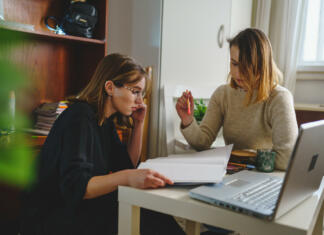 Kaj je „tutorstvo“? two women young caucasian female student sitting at home with her mentor teacher looking to the notebook explaining lesson study preparing for exam learning education concept real people copy space