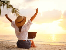 Work from anywhere. Rear view of young woman, female freelancer in straw hat working on laptop, keeping arms raised and cellebrating success while sitting on the tropical sandy beach at sunset