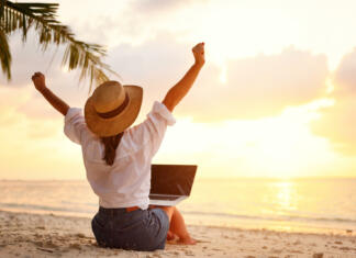 Delo v tujini Work from anywhere. Rear view of young woman, female freelancer in straw hat working on laptop, keeping arms raised and cellebrating success while sitting on the tropical sandy beach at sunset