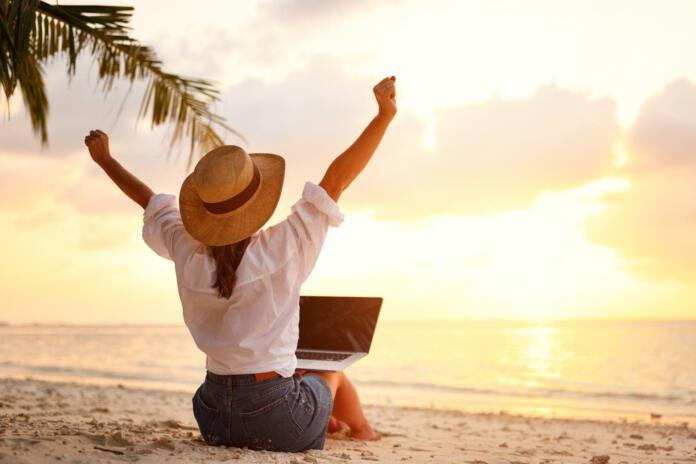 work-from-anywhere-rear-view-of-young-woman-female-freelancer-in-straw-hat-working-on-laptop-keeping-arms-raised-and-cellebrating-success-while-sitting-on-the-tropical-sandy-beach-at-sunset-stockpack-istock Work from anywhere. Rear view of young woman, female freelancer in straw hat working on laptop, keeping arms raised and cellebrating success while sitting on the tropical sandy beach at sunset