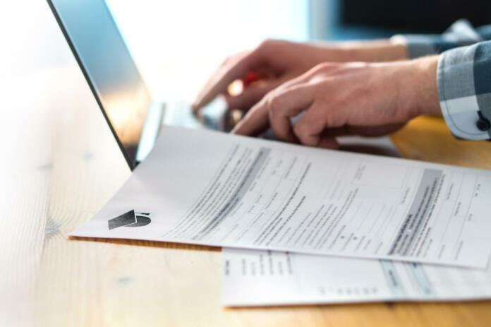 Young man writing college or university application form with laptop. Student applying to school. Scholarship document, admission paper or letter on table. Typing email. Education and communication.