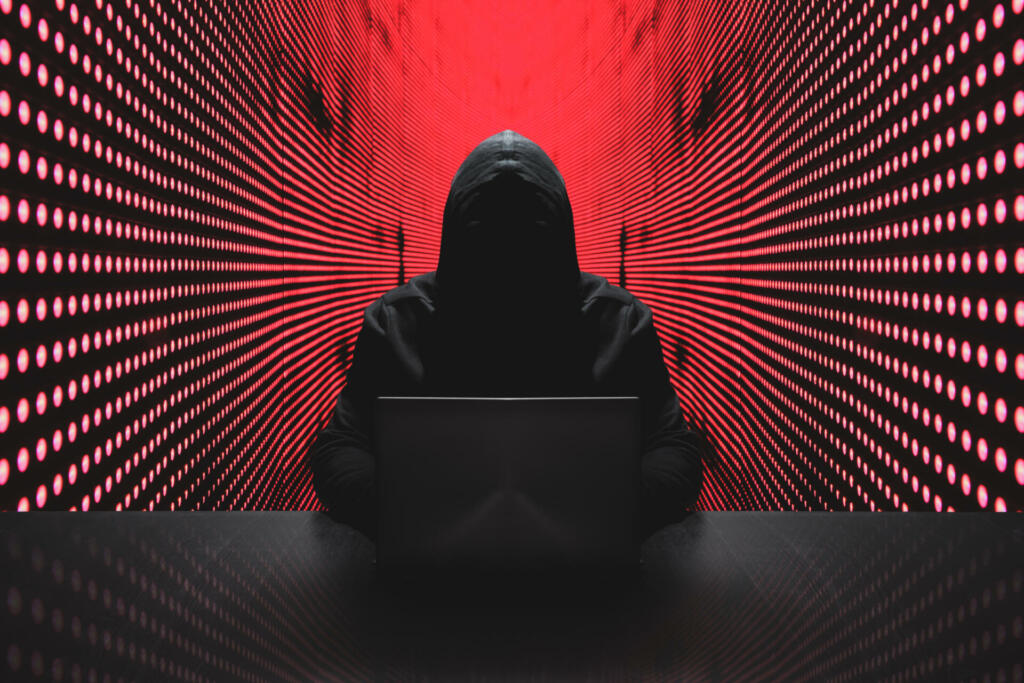 Anonymous hacker in front of his computer with red light wall backgroundAnonymous hacker in a black hoody with laptop in front of a code background with binary streams cyber security concept