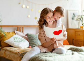 Materinski dan Cheerful mother hugging son and reading handmade greeting card with heart while resting on bed during holiday celebration mothers day  at home