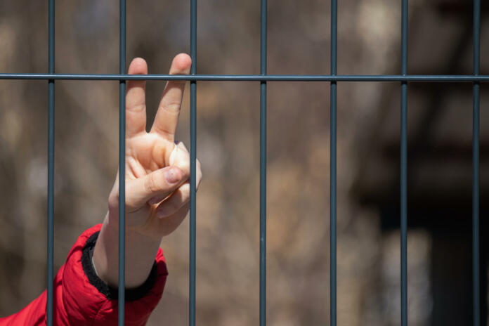 Child refugee showing peace sign behind a metal fence. Social problem of war migrants.