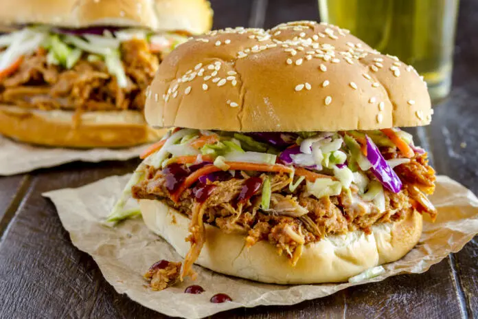 Close up of pulled pork barbeque sandwich with coleslaw sitting on wooden table with glass of beer