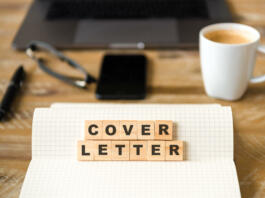 Closeup on notebook over wood table background, focus on wooden blocks with letters making COVER LETTER words. Business concept image. Laptop, glasses, pen and mobile phone in a defocused background.