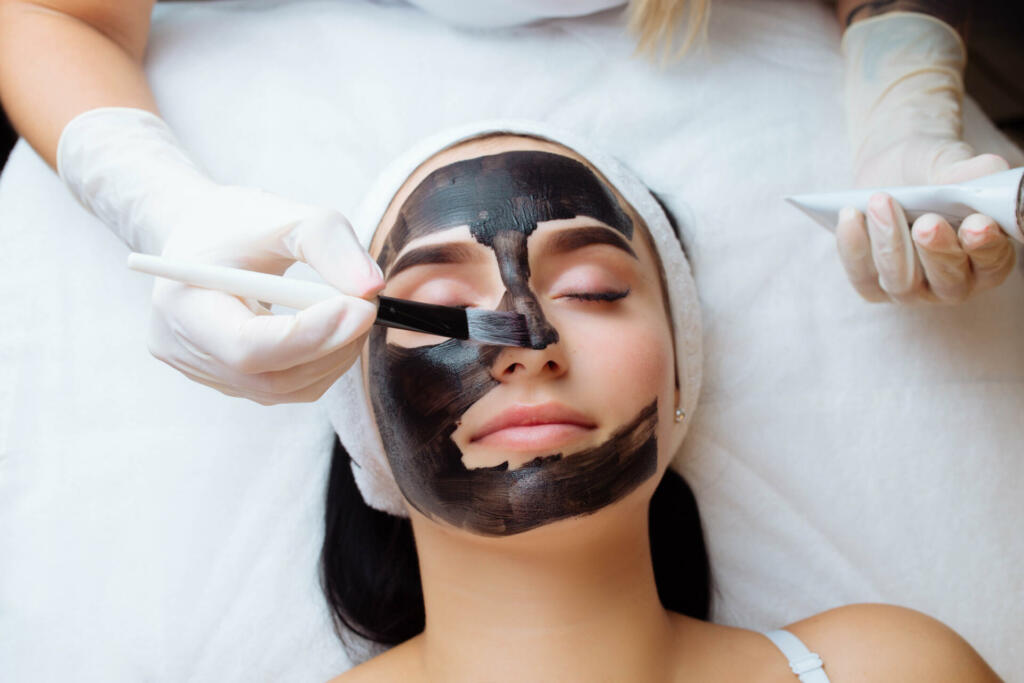 cosmetologist applying black mask on pretty woman face wearing black gloves, gorgeous woman in spa having facial procedures.