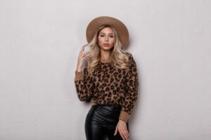 Elegant glamorous young woman with beautiful makeup with curly hair in a luxurious hat in a leopard sweater in fashionable black leather pants posing in a room near a white wall. Sexy attractive girl.