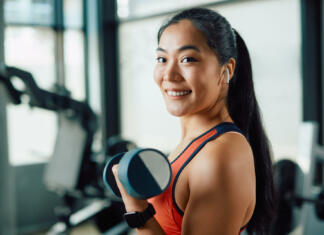 Postani fit: naboljše vaje za hrbtne mišice in lepo držo Happy Asian sportswoman lifting dumbbells during strength training in a gym and looking at camera.