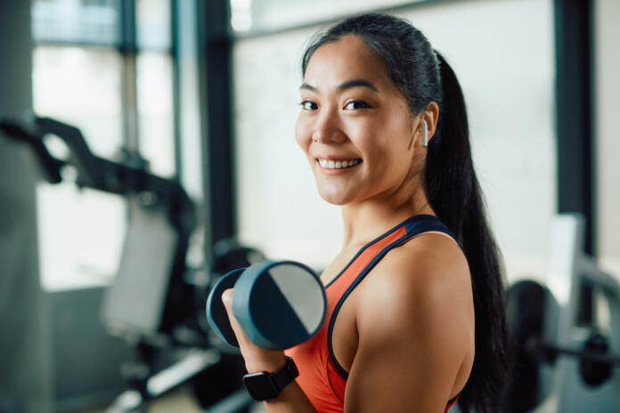 Happy Asian sportswoman lifting dumbbells during strength training in a gym and looking at camera.