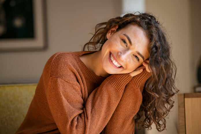 Happy young woman sitting on sofa at home and looking at camera. Portrait of comfortable woman in winter clothes relaxing on armchair. Portrait of beautiful girl smiling and relaxing during autumn.