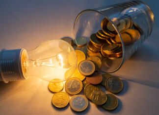 Bo vladin ukrep nižanja DDV pomagal pri energetski draginji? Incandescent light bulb turned on, with jar full of coins next to it, on wooden surface. Increased costs. Savings. Energy tariffs.