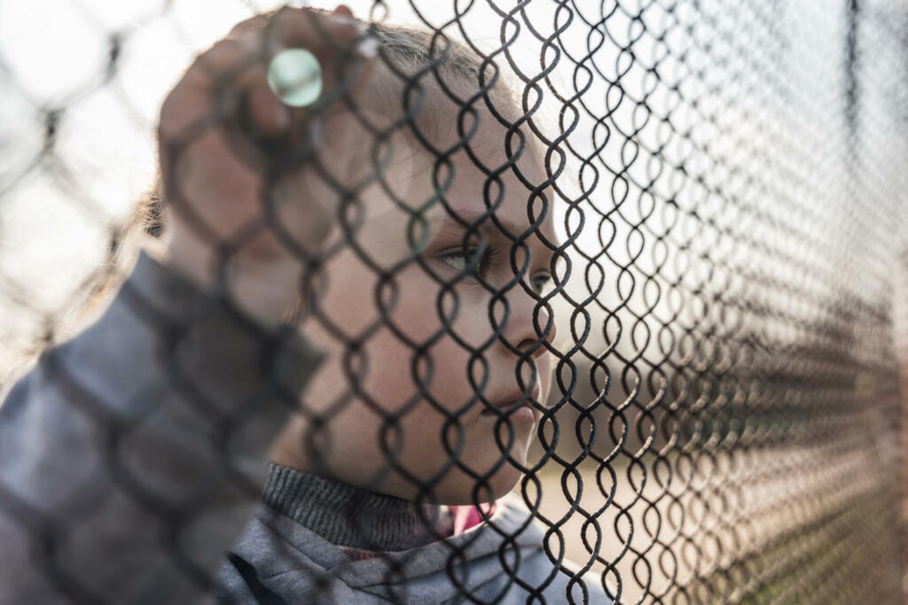 Little girl with a sad look behind a metal fence, social problems, raising children in orphanages.