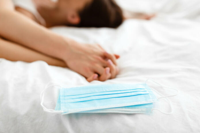 Love And Dating During Covid-19 Pandemic. Protective Face Masks Lying Near Passionate Couple Holding Hands Having Sex In Bed Indoors. Conceptual Shot. Selective Focus, Shallow Depth