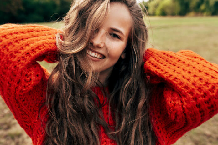 Outdoor portrait of a smiling young woman wearing a knitted orange sweater posing on nature background. The beautiful female has a joyful expression, resting in the park.