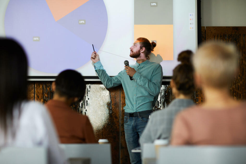 Portrait of contemporary bearded man giving presentation standing by projector screen in lecture hall, copy space