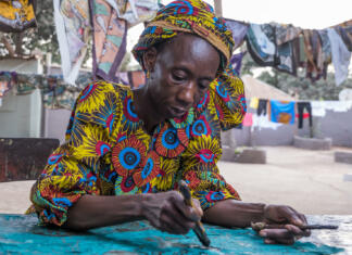 Študent v Gambiji: perspektiva prostovoljke Zymryte Ademaj Serekunda, The Gambia - June 22, 2019: Old woman painting fabrics in a traditional way in a popular neighborhood of the region