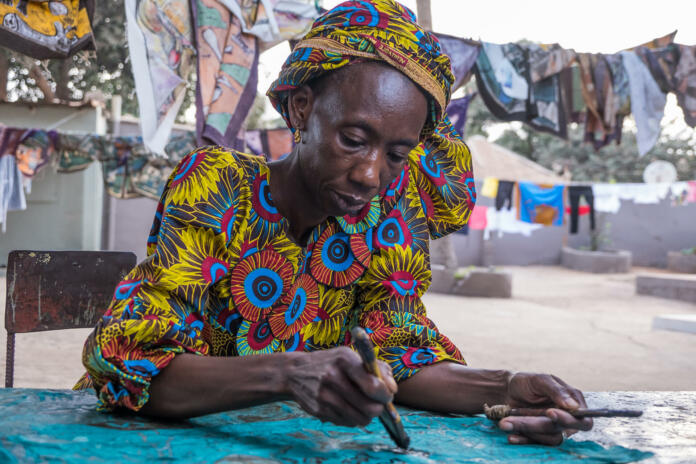 Serekunda, The Gambia - June 22, 2019: Old woman painting fabrics in a traditional way in a popular neighborhood of the region