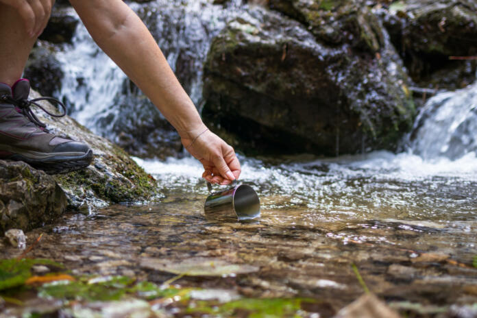Thirsty hiker scoops fresh water into cup from stream in mountains. Refreshment during hiking in nature