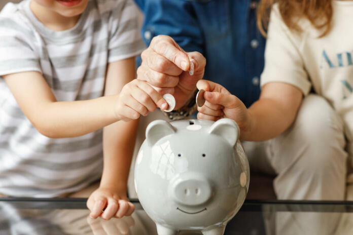 Unrecognizable senior man and kids putting coins into piggy bank on table at home together