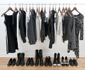 Women's clothing hanging from a metal bar and shoes on the hardwood floor beneath them.  The clothes and shoes are primarily black with some white and gray.  A white wall is in the background.