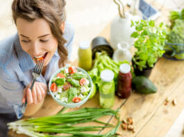 Young and happy woman eating healthy salad sitting on the table with green fresh ingredients indoors