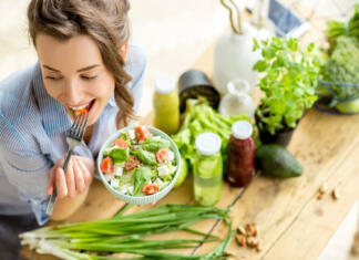 Young and happy woman eating healthy salad sitting on the table with green fresh ingredients indoors