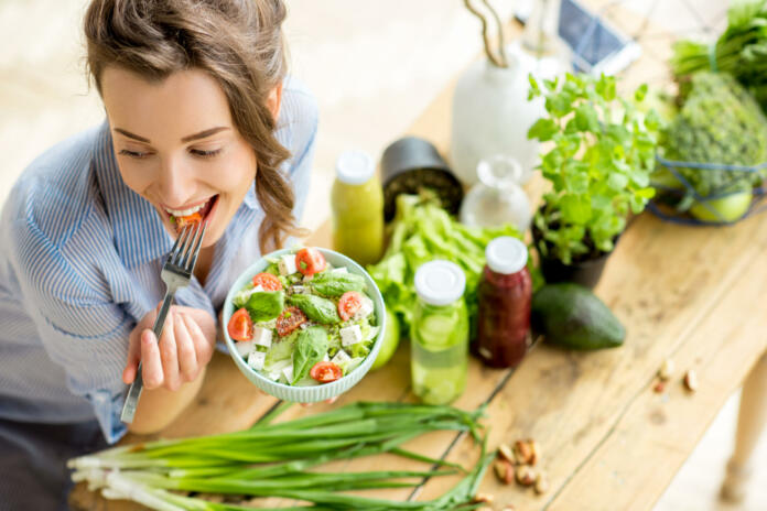 Young and happy woman eating healthy salad sitting on the table with green fresh ingredients indoors