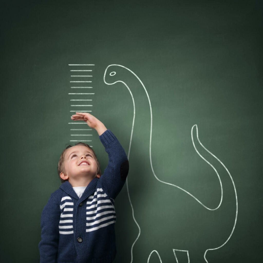 Young boy measuring his growth in height against a blackboard with chalk dinosaur scale