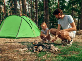 A young father and his daughter sit by a campfire near a tent and grill marshmallows during the weekend in a pine forest. Camping, recreation, hiking.