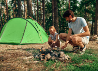 Kaj vse potrebuješ za kampiranje v naravi? Pripravili smo ti seznam! A young father and his daughter sit by a campfire near a tent and grill marshmallows during the weekend in a pine forest. Camping, recreation, hiking.