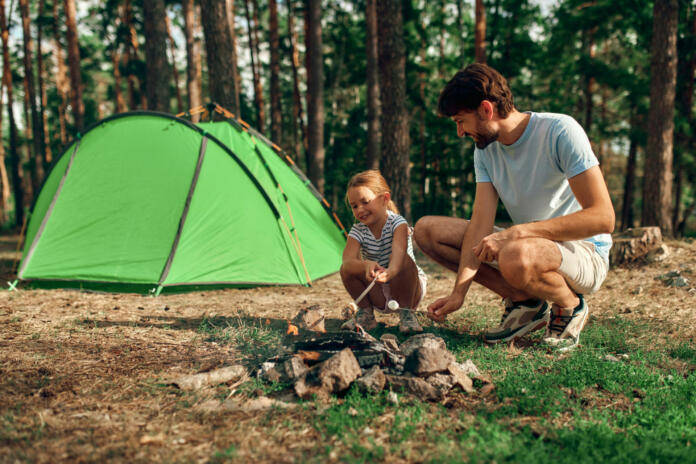 A young father and his daughter sit by a campfire near a tent and grill marshmallows during the weekend in a pine forest. Camping, recreation, hiking.