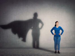Brave woman keeps arms on hips, smiling confident, casting a superhero with cape shadow on the wall. Ambition and business success concept. Leadership hero power, motivation and inner strength symbol.
