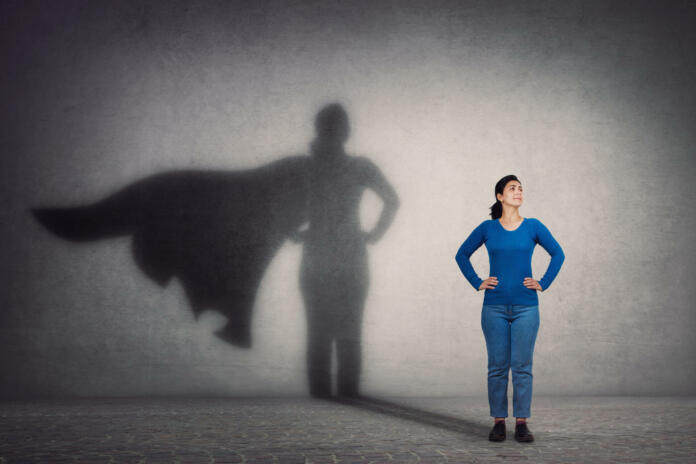 Brave woman keeps arms on hips, smiling confident, casting a superhero with cape shadow on the wall. Ambition and business success concept. Leadership hero power, motivation and inner strength symbol.