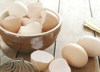 12 stvari, za kaj uporabimo jajčne lupine? (Posodobljeno 2022) Egg shells in ceramic bowl. Some eggs, egg beater and table cloth next to it. All on wooden table.