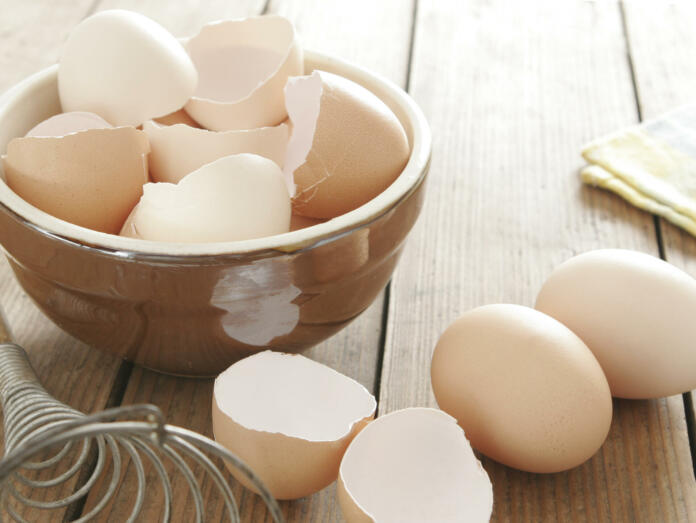 Egg shells in ceramic bowl. Some eggs, egg beater and table cloth next to it. All on wooden table.