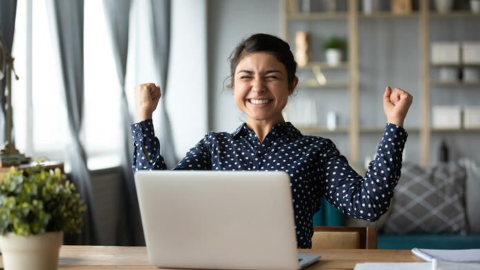 Euphoric young indian girl student winner celebrate victory triumph sit at home desk with laptop computer win online fortune feel excited get new job opportunity good exam result great news concept