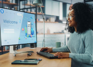 Kakšen računalnik rabim za grafični dizajn? Handsome Black Female is Using Desktop Computer that Shows Welcome Page of a Popular Social Network. Freelance Woman Working Over the Internet as a Social Network Marketer.
