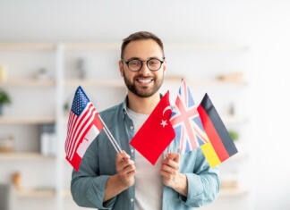 Zgodbe uspehov, povezane s tujimi jeziki Handsome young guy holding bunch of diverse flags and smiling at camera indoors. Happy millennial man recommending foreign languages school, emigrating abroad. Modern education and student exchange